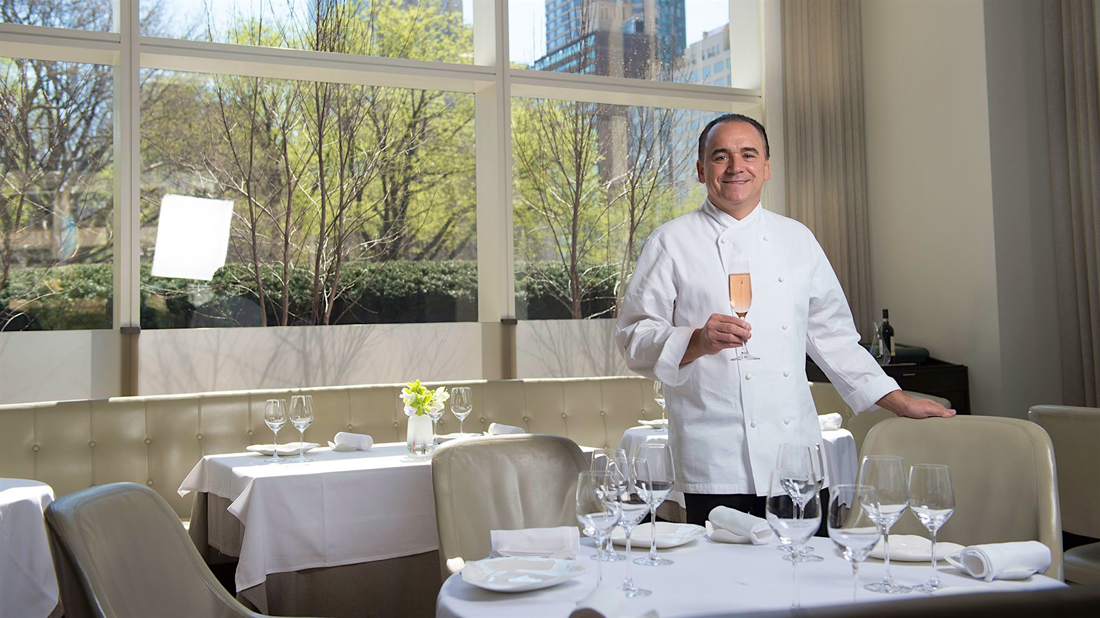 Jean-Georges Vongerichten with a glass of sparkling wine in the dining room of Jean-Georges in New York, with cream-colored leather chairs and banquettes and a large window looking onto the west side of Manhattan